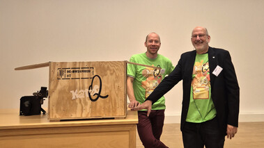 Zwei Männer stehen bei einer Preisverleihung neben einer Holzkiste mit KatzeQ-Logo.